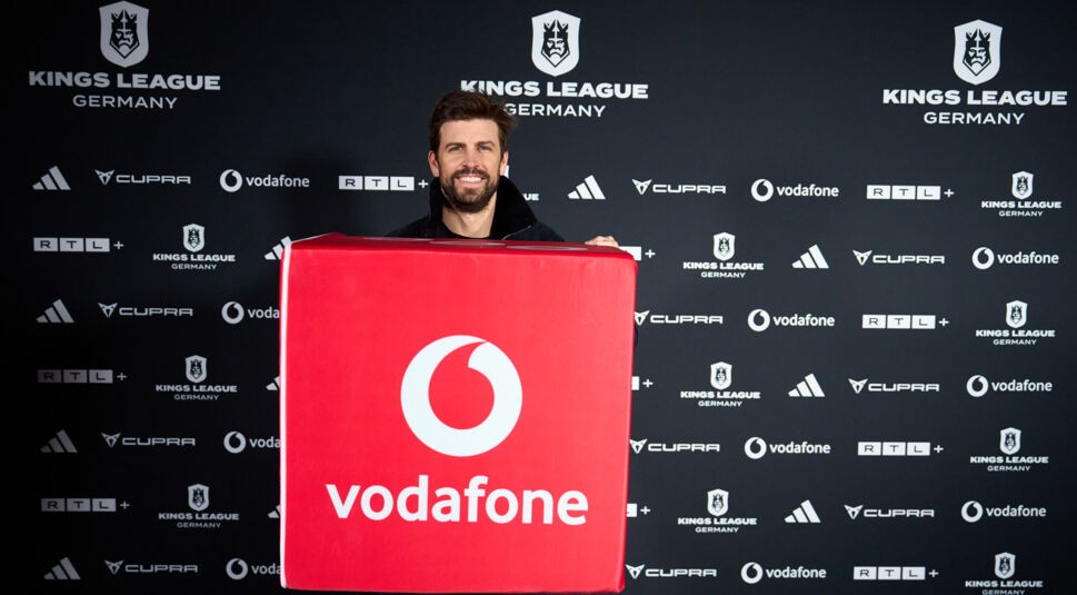 Auf dem Bild zur Kings League Germany steht Gerard Piqué lächelnd vor einer Sponsorenwand und hält eine große, rote Box mit prominentem Vodafone-Logo in den Händen. Die Wand im Hintergrund zeigt Logos von RTL+, Adidas, Cupra und der Kings League Germany. Die Szene wirkt dynamisch und unterstreicht die Partnerschaft mit Vodafone.