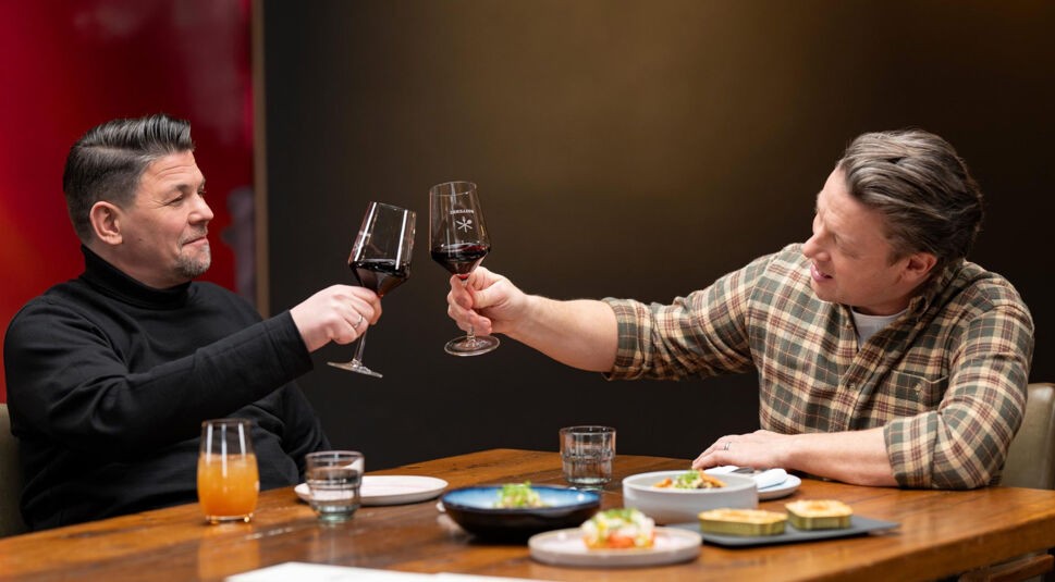 In dieser Szene aus Kitchen Impossible Staffel 10 stoßen Tim Mälzer und Jamie Oliver mit Rotweingläsern an. 