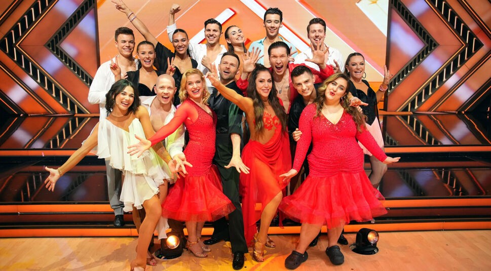 Auf dem Bild zur Let's Dance 2025 Show 7 sind alle Tanzpaare fröhlich und ausgelassen vor der Bühne versammelt. In farbenfrohen Outfits, überwiegend in Rot, posieren sie in einer lebendigen Formation. Die Stimmung wirkt feierlich und energiegeladen.