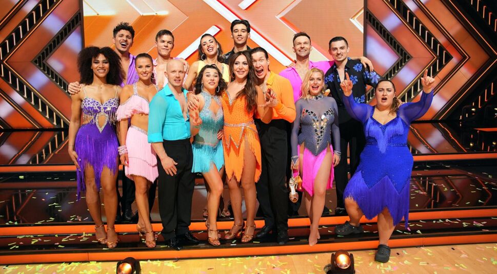 Auf dem Bild zur Let's Dance 2025 Show 8 posiert die gesamte Tanz-Crew freudestrahlend auf der Showbühne. Die Teilnehmer:innen tragen bunte, glitzernde Tanzkostüme in Lila, Blau, Orange und Pink. Die Stimmung wirkt ausgelassen und feierlich, bunte Konfetti liegen auf dem Boden. Im Hintergrund ist das auffällige Bühnendesign von RTL zu sehen.