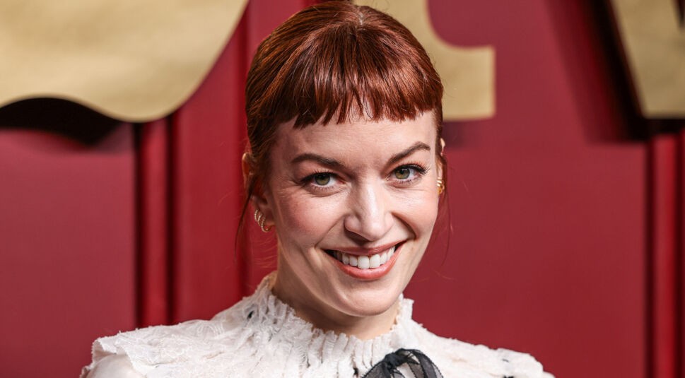 Auf dem Bild zur Nur für Dein Leben Netflix-Serie ist Britt Lower vor einem roten Hintergrund mit goldenen Buchstaben zu sehen. Sie trägt eine elegante weiße Spitzenbluse mit feinen Details und lächelt offen in die Kamera. Ihr rötlich-braunes Haar ist streng zurückgebunden, ihr Blick wirkt freundlich und herzlich, was der Szene eine charmante und einladende Stimmung verleiht.