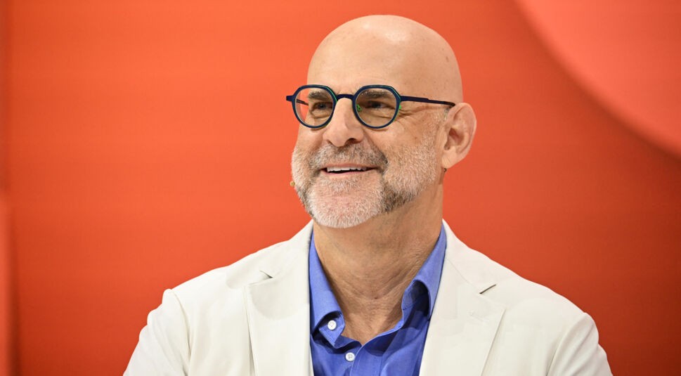 Auf dem Bild zur Nur für Dein Leben Netflix-Serie ist der Autor Harlan Coben vor einem kräftig orangenen Hintergrund zu sehen. Er trägt ein helles Sakko über einem blauen Hemd und eine runde Brille mit dunklem Rahmen. Sein Gesichtsausdruck ist freundlich und offen, er lächelt leicht zur Seite. Die Szene wirkt professionell und optimistisch.