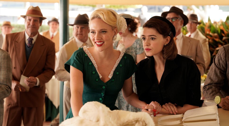 Auf diesem Bild zur On Swift Horses-Kritik sind Gail, gespielt von Kat Cunning, und Muriel, dargestellt von Daisy Edgar-Jones, in einer eleganten Szene der 1950er Jahre zu sehen. Gail trägt ein grünes Kleid mit weißen Details und einem auffälligen Haarschmuck, während Muriel in Schwarz gekleidet ist. Beide wirken gespannt und lebendig, umgeben von elegant gekleideten Menschen in nostalgischer Atmosphäre.