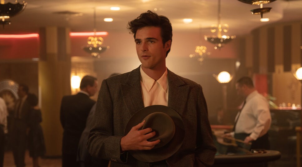 Im Bild zur On Swift Horses-Kritik ist Julius, dargestellt von Jacob Elordi, in einem stilvollen Casino-Ambiente der 1950er Jahre zu sehen. Er trägt ein kariertes Sakko und hält lässig einen Hut in der Hand. Mit selbstbewusstem Blick steht er im Halbdunkel des Raumes, während hinter ihm Menschen an Spieltischen agieren. Die Szene wirkt geheimnisvoll und atmosphärisch dicht.