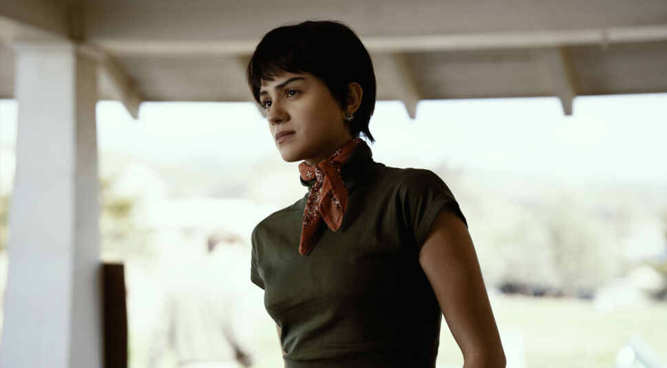 Das Bild zur On Swift Horses-Kritik zeigt Sandra, gespielt von Sasha Calle, in einem nachdenklichen Moment. Sie trägt ein eng anliegendes olivgrünes Shirt und ein rotes Halstuch, das ihren rebellischen Look unterstreicht. Mit festem Blick schaut sie zur Seite, im Hintergrund verschwimmen helle Landschaft und Personen in der Unschärfe. Die Szene vermittelt Entschlossenheit und innere Stärke.