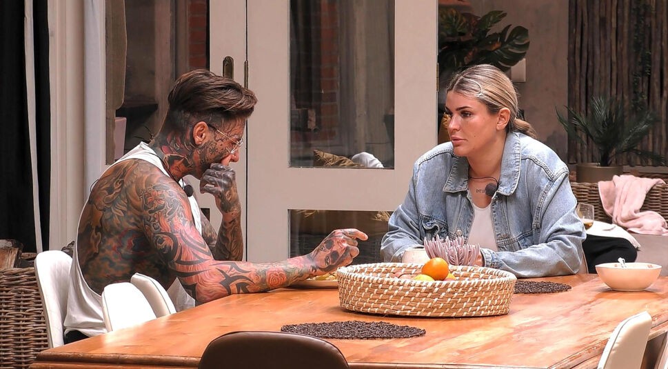 In einer Szene aus Staffel 2 von „Prominent getrennt“ (2023) sitzen Nikola Glumac und Gloria Glumac an einem Holztisch und führen ein ernstes Gespräch. Nikola, stark tätowiert und im weißen Tanktop, gestikuliert nachdenklich, während Gloria ihm ruhig gegenübersitzt. Das Bild zeigt die emotionalen Momente der späteren „Prominent getrennt-Gewinner:innen“.