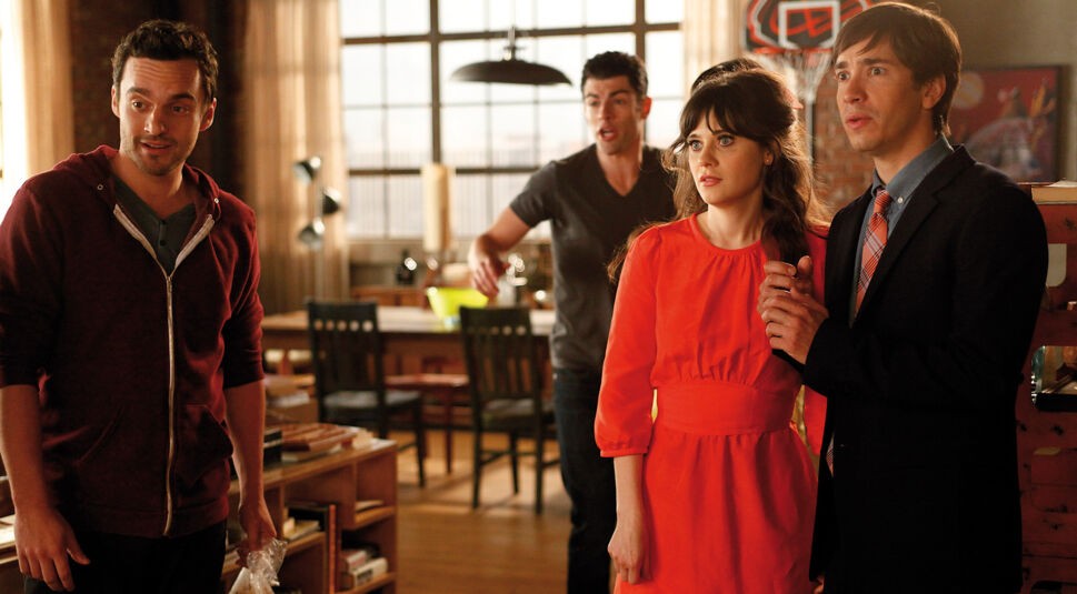 Auf dem Bild in dem Artikel zu den Serien wie The Big Bang Theory ist eine Szene aus der Serie New Girl zu sehen. Vier Personen stehen in einem gemütlich eingerichteten Loft, die Stimmung wirkt angespannt, aber humorvoll. Im Hintergrund ist ein Esstisch mit Stühlen zu erkennen, das Licht fällt warm durch die großen Fenster.