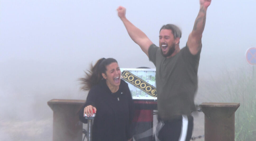 Auf dem Bild in dem Artikel zu den Sommerhaus der Stars-Gewinner:innen jubeln Elena Miras und Mike Heiter aus Staffel 4 ausgelassen über ihren Sieg. Im Hintergrund ist ein silberner Geldkoffer mit der Aufschrift „50.000 €“ zu sehen. Der Nebel und die natürliche Umgebung verleihen der Szene eine dramatische, aber triumphale Stimmung.