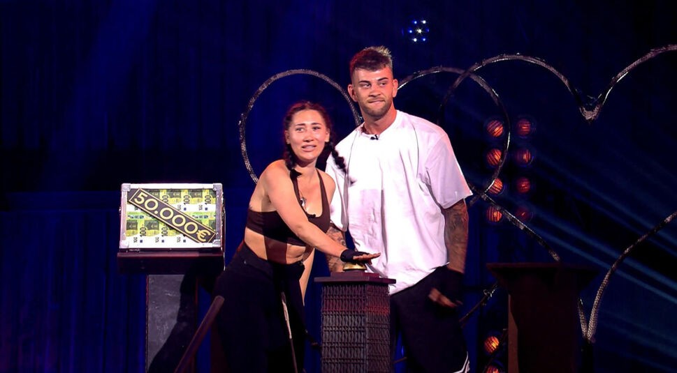 Auf dem Bild in dem Artikel zu den Sommerhaus der Stars-Gewinner:innen stehen Samira und Serkan Yavuz aus Staffel 8 beim Finalspiel vor einem Geldkoffer mit der Aufschrift „50.000 €“. Samira drückt mit entschlossenem Blick den Buzzer, Serkan steht dicht neben ihr. Hinter ihnen stehen zwei Herzformen, die Bühne ist in dramatisches Licht getaucht.