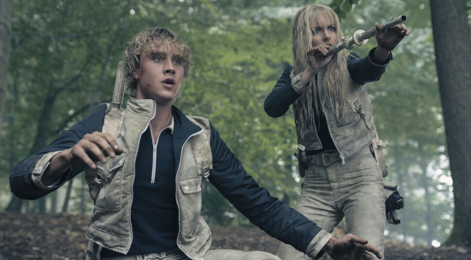 Auf dem Bild zu Tribute von Panem 6 befinden sich Mckenna Grace als Maysilee Donner und Joseph Zada als junger Haymitch Abernathy in einer angespannten Kampfszene im Wald. Beide tragen schmutzige Tribute-Uniformen, Maysilee zielt mit einer improvisierten Waffe, während Haymitch alarmiert wirkt. Die düstere Waldkulisse verstärkt die bedrohliche Stimmung der Arena.