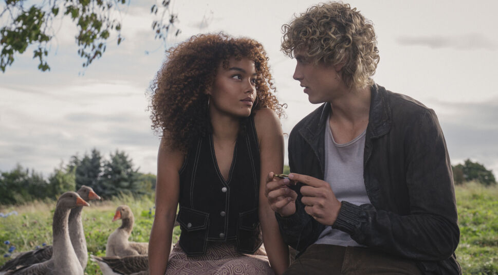 Auf dem Bild zu Tribute von Panem 6 sind Whitney Peak als Lenore Dove und Joseph Zada als junger Haymitch Abernathy in einer ruhigen, nachdenklichen Szene zu sehen. Die beiden sitzen auf einer Wiese, umgeben von Gänsen, und blicken sich intensiv an. Die ländliche Umgebung wirkt friedlich und steht im Kontrast zur angespannten Atmosphäre zwischen den Charakteren.