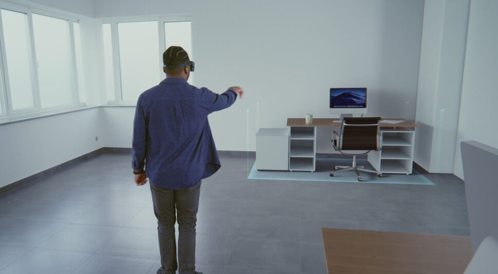 Das symbolische Bild zur Frage „Was ist Mixed Reality“ zeigt einen Mann mit Mixed-Reality-Brille in einem leeren Raum. Er deutet auf ein virtuelles Schreibtisch-Setup mit Monitor, Bürostuhl und Tasse, das holografisch vor ihm erscheint. Die Szene vermittelt den Einsatz von Mixed Reality zur Visualisierung und Interaktion mit digitalen Arbeitsumgebungen in realen Räumen.