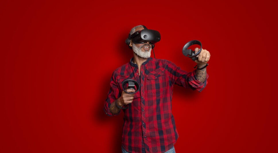 Ein älterer Mann mit grauem Bart und tätowiertem Arm trägt eine Virtual-Reality-Brille und hält zwei Controller in den Händen. Er steht vor einem knallroten Hintergrund und wirkt begeistert und konzentriert. Das Bild visualisiert auf moderne und dynamische Weise die Frage: Was ist Virtual Reality?