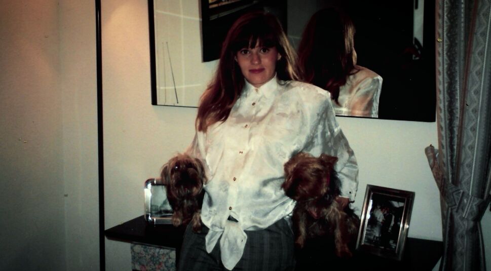 Angi - (K)eine perfekte Mörderin: Eine junge Frau mit weißer Bluse hält zwei kleine Hunde im Arm und steht vor einem Spiegel und einer Kommode mit Fotos.