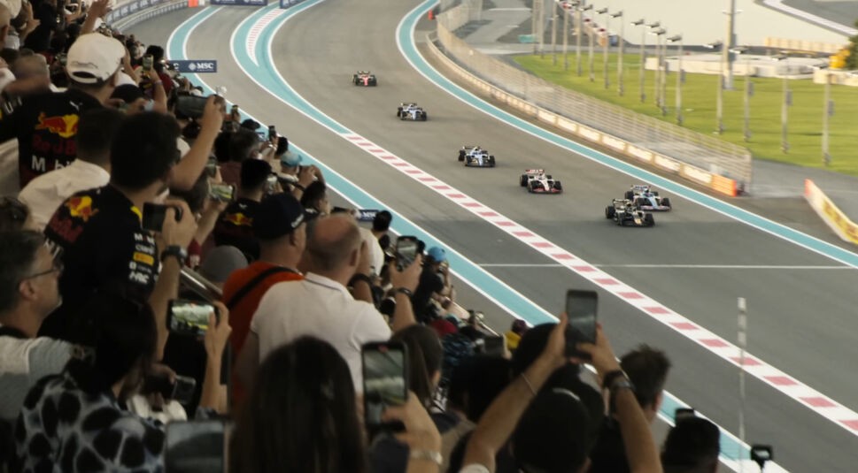 Bild aus dem Film F1: Formel-1-Autos rasen auf der Strecke, während jubelnde Zuschauer in der Tribüne das Rennen mit ihren Smartphones festhalten.