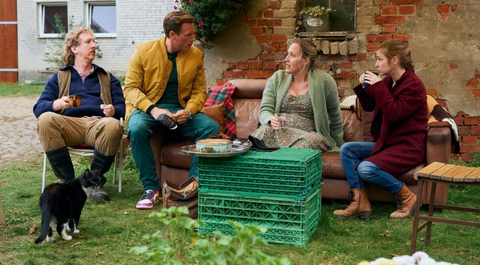 Uke Bosse, Patrick Kalupa, Luise Schnittert und Josefine Preuß sitzen in einer Szene von Dr. Nice während einer Teepause im Garten eines Bauernhofes.