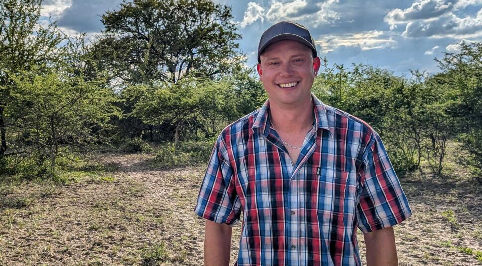 Björn, Kandidat der RTL-Sendung „Bauer sucht Frau international Staffel 7“, steht lachend auf trockenem Boden in der savannenartigen Landschaft Namibias. Er trägt ein kariertes Hemd in Blau-Rot und eine dunkle Cap. Die Sonne scheint durch eine aufgelockerte Wolkendecke, während Büsche und Bäume im Hintergrund das ländliche Setting betonen.