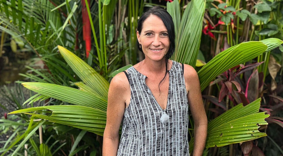 Carmen, eine Teilnehmerin der RTL-Sendung „Bauer sucht Frau international Staffel 7“, steht lächelnd vor tropischen Pflanzen auf Bali. Sie trägt ein ärmelloses Oberteil mit schwarz-weißem Muster. Die Kulisse mit großen, grünen Blättern und exotischen Farben unterstreicht die tropische Stimmung des Settings und betont Carmens offene Ausstrahlung.