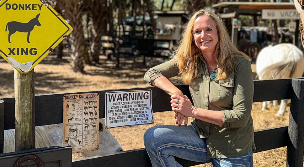 Iris, Kandidatin der RTL-Sendung „Bauer sucht Frau international Staffel 7“, lehnt entspannt an einem Zaun auf ihrer Farm in Florida. Im Hintergrund sind ein Esel, ein Pferd und mehrere Hinweisschilder zu sehen, darunter ein gelbes „Donkey Xing“-Schild. Sie trägt Jeans und ein grünes Hemd und lächelt freundlich in die Kamera bei sonnigem Wetter.