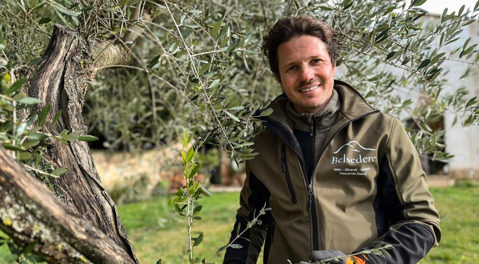 Martin, Teilnehmer der RTL-Sendung „Bauer sucht Frau international Staffel 7“, steht zwischen den Zweigen eines Olivenbaums auf seinem Hof in der Toskana. Er trägt eine Jacke mit der Aufschrift „Belsedere“ und lächelt freundlich. Die Szene wirkt ruhig und naturverbunden, mit grünem Rasen und traditioneller Architektur im Hintergrund.
