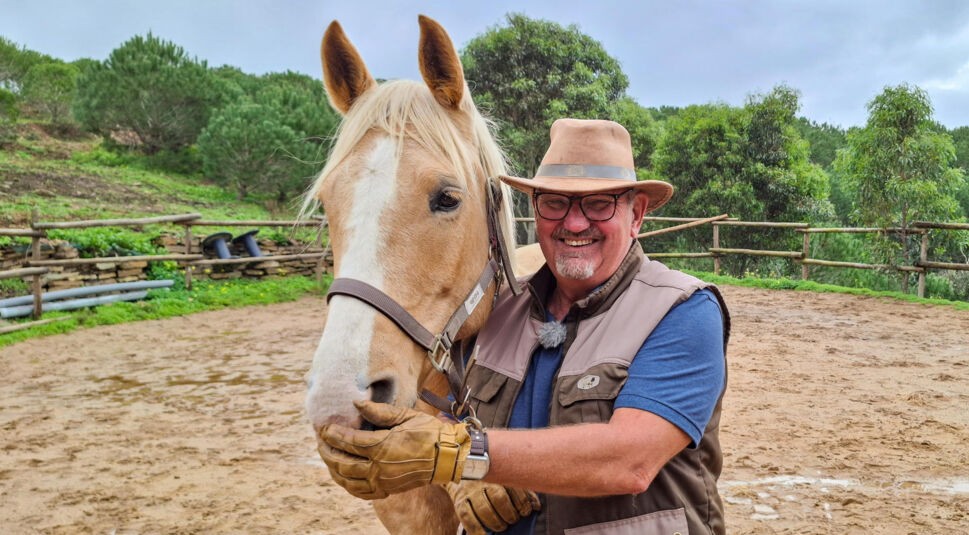 Olaf, Kandidat der RTL-Sendung „Bauer sucht Frau international Staffel 7“, steht auf seiner Pferdekoppel in Portugal. Er trägt einen beigen Hut, Handschuhe und eine Outdoor-Weste und lächelt zufrieden in die Kamera. Neben ihm steht ein hellbraunes Pferd mit weißer Blesse, das er liebevoll am Maul hält. Im Hintergrund sind grüne Bäume und ein rustikaler Zaun zu sehen.