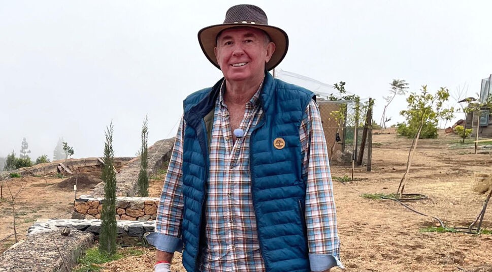 Roger, Kandidat der RTL-Sendung „Bauer sucht Frau international Staffel 7“, steht gut gelaunt auf seinem Hof auf Teneriffa. Er trägt ein kariertes Hemd, eine blaue Steppweste und einen braunen Hut. Die Landschaft ist trocken mit Steinmauern, jungen Bäumen und leichter Hanglage, während im Hintergrund Nebel aufzieht – typisch für die Höhenlagen der Kanareninsel.