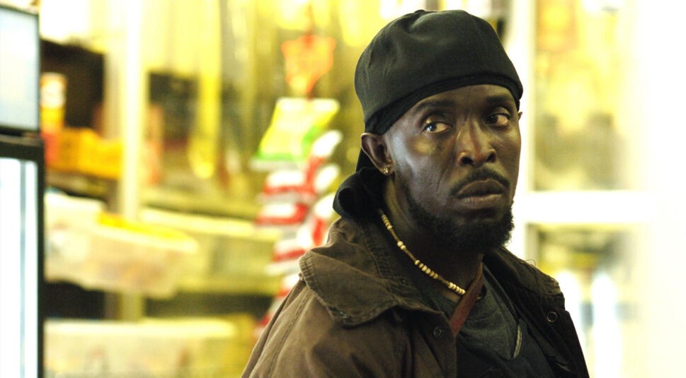Auf diesem Bild zur „The Wire“-Serie ist Omar Little zu sehen, gespielt von Michael Kenneth Williams. Er steht in einem kleinen Laden, trägt eine dunkle Jacke, ein Bandana und eine Perlenkette. Sein ernster, misstrauischer Blick unterstreicht die angespannte Stimmung der Szene, typisch für die düstere Atmosphäre der The Wire-Serie.