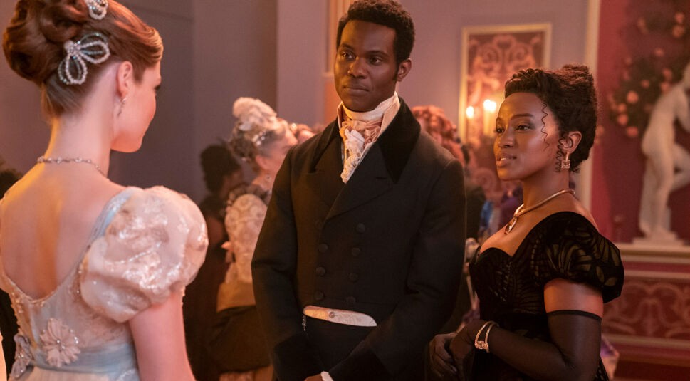 Auf dem Bild in dem Artikel zu Bridgerton Staffel 5 steht Francesca Bridgerton mit dem Rücken zur Kamera und spricht mit John Stirling und Michaela. Francesca trägt ein helles Ballkleid, John ist in einem dunklen Frack mit hellem Halstuch gekleidet, während Michaela in einem eleganten schwarzen Kleid mit aufwendigem Muster und Schmuckstücken selbstbewusst wirkt. 