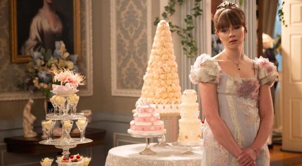 Auf dem Bild in dem Artikel zu Bridgerton Staffel 5 steht Eloise in einem pastellfarbenen Ballkleid mit floralen Verzierungen nachdenklich neben einem festlich gedeckten Tisch voller aufwendig verzierter Torten und Desserts.