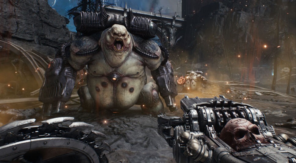 In dieser Szene aus Doom: The Dark Ages Tipps und Tricks im Guide greift ein riesiger, fetter Dämon mit wuchtigen Kanonenarmen frontal an. Der Spieler hält eine brutale Waffe mit Totenschädel-Design und rotierenden Elementen im Vordergrund. Umgeben von glühenden Partikeln und brennender Umgebung herrscht eine aggressive, düstere Stimmung auf dem zerstörten Schlachtfeld.