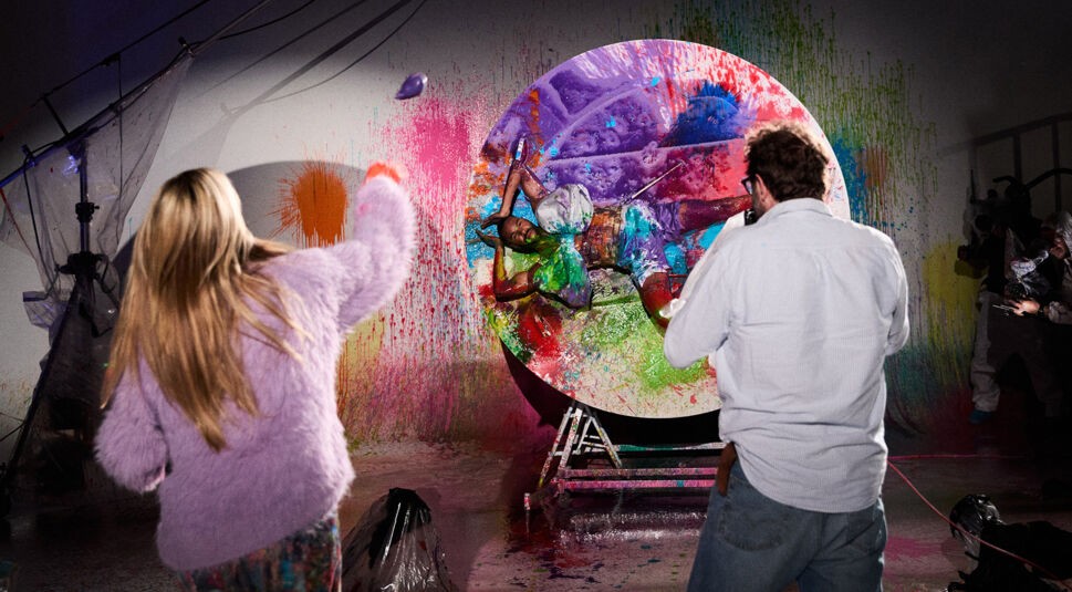 Auf dem Bild zu Folge 19 von GNTM 2025 wirft Heidi Klum eine Farbbombe auf einen ihrer Kandidaten, der an einer Drehscheibe versucht, für ein perfektes Foto zu posieren. Rechts neben ihr steht der Fotograf Christian Anwander.