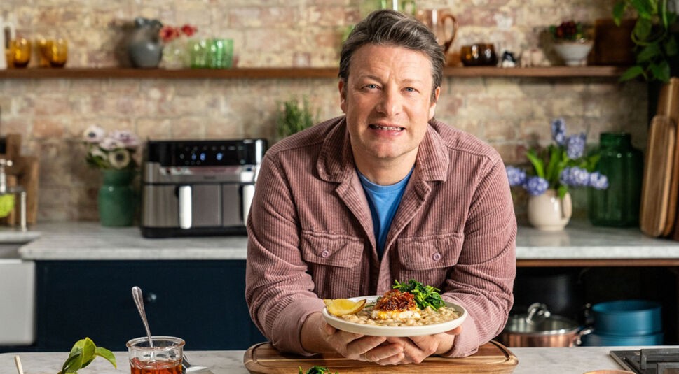 In dem Artikel zu den kostenlosen Jamie Oliver Folgen zu seinem 50. Geburtstag ist Jamie Oliver in einer gemütlichen Studioküche zu sehen. Er trägt eine rosa Cordjacke, blickt freundlich in die Kamera und präsentiert ein liebevoll angerichtetes Tellergericht mit Fisch, Bohnen und Gemüse.