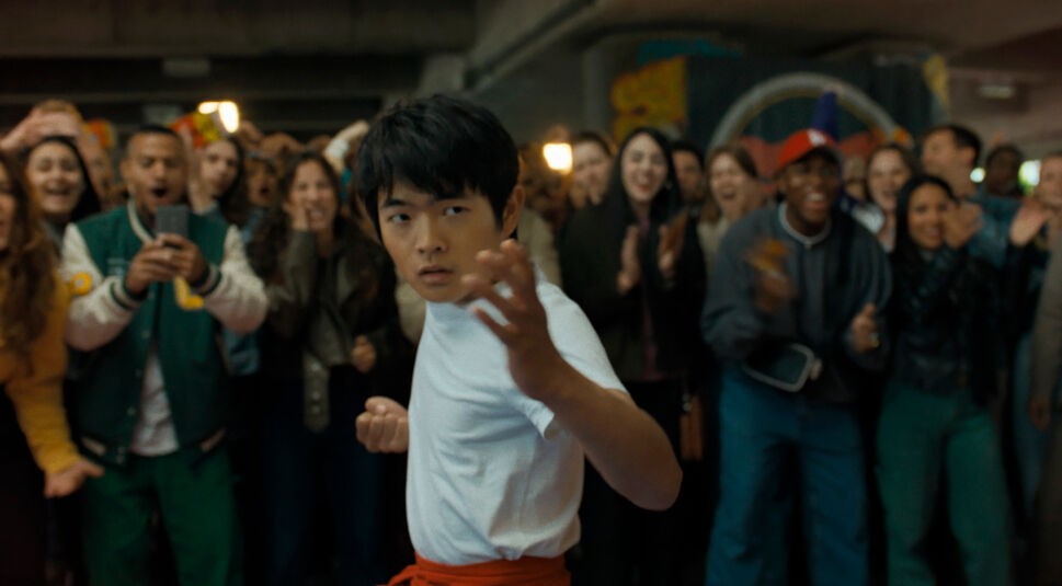 Ben Wang steht in dieser Szene aus Karate Kid: Legends kampfbereit in einem weißen Shirt mit rotem Gürtel in einer Menschenmenge. Mit entschlossenem Blick und fokussierter Haltung signalisiert er Bereitschaft und Mut. Hinter ihm jubelt eine diverse Gruppe junger Zuschauer – ein emotionaler Moment für die Karate Kid: Legends Kritik.