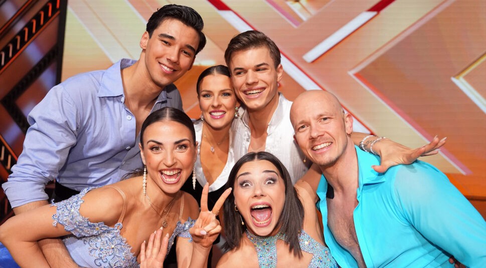 Auf diesem Bild zum Lets Dance 2025 Finale sind die Finalist:innen der Staffel in ausgelassener Stimmung zu sehen. In der vorderen Reihe lachen und posieren die Profitänzerinnen und Promis Arm in Arm, während der bunte Hintergrund der RTL-Show für festliche Atmosphäre sorgt. Alle tragen glamouröse Outfits, die ihre Auftritte widerspiegeln.
