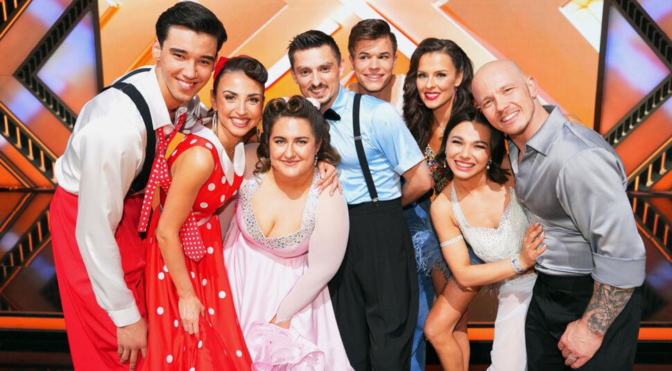 Auf dem Bild zur Let's Dance 2025 Show 11 sind acht Teilnehmende der Tanzshow zu sehen. Die Gruppe posiert lächelnd vor einem farbenfrohen Bühnenhintergrund. Die Outfits reichen von glitzernden Tanzkleidern bis zu Retro-Kostümen mit Hosenträgern, was auf unterschiedliche Tanzstile in der Show hindeutet.
