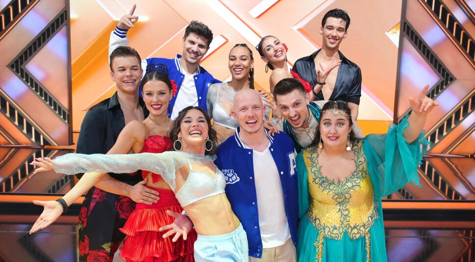 Auf dem Bild zu Lets Dance 2025 sind zehn Tänzer:inne fröhlich und energiegeladen vor einem leuchtenden Bühnenhintergrund zu sehen. Alle tragen auffällige Kostüme – von glitzernden Tops über bunte Tanzkleider bis zu sportlichen Collegejacken. Die Stimmung wirkt ausgelassen und sie lachen direkt in die Kamera.
