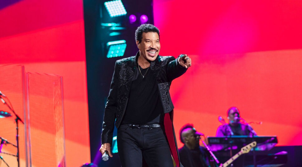 Im Artikel zu den Lionel Richie Tickets ist Lionel Richie auf diesem Bild mitten in der Interaktion mit seinem Publikum zu sehen. Er zeigt lachend und selbstbewusst in den Zuschauerraum, während er ein Mikrofon in der linken Hand hält.