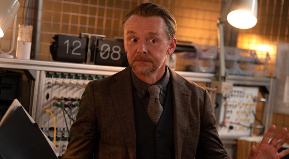 Im Bild zur Mission: Impossible 8 Kritik ist Simon Pegg als Benji Dunn in einem Technikraum zu sehen. Er trägt einen braunen Anzug mit Krawatte und schaut nervös zur Seite. Hinter ihm befindet sich ein mit Kabeln und Schaltern gespicktes Kontrollpanel, das auf eine angespannt-technologische Situation im IMF-Team hinweist.