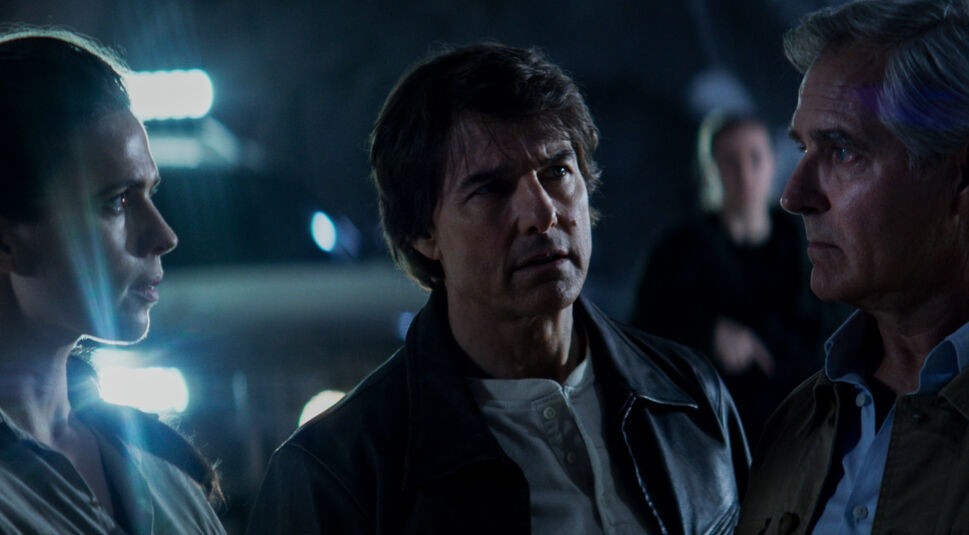 Im Bild zur Mission: Impossible 8 Kritik stehen Hayley Atwell als Grace, Tom Cruise als Ethan Hunt und Henry Czerny als Kittridge angespannt beieinander. Die Szene spielt in einer düsteren Umgebung mit starker Gegenlichtbeleuchtung, was die Dramatik und Ernsthaftigkeit ihrer Besprechung unterstreicht. Ihre Blicke sind konzentriert, die Stimmung wirkt angespannt und entscheidend.