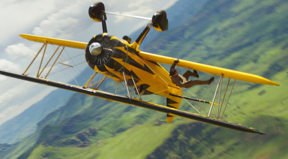 Im Bild zur Mission: Impossible 8 Kritik ist Tom Cruise als Ethan Hunt in einer waghalsigen Actionszene zu sehen. Er hängt kopfüber an einem gelb-schwarzen Doppeldeckerflugzeug, das sich in rasanter Schräglage über eine grüne Hügellandschaft bewegt. Die Szene vermittelt Spannung, Risiko und den für die Filmreihe typischen Adrenalinrausch.