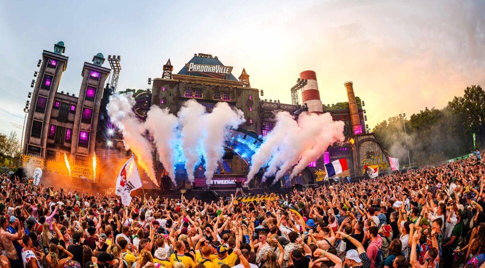 Dieses Bild zum Thema parookaville tickets zeigt die energiegeladene Stimmung auf dem Parookaville-Festival am Tag. Tausende Menschen tanzen vor der gigantischen Hauptbühne, während Rauchfontänen aufsteigen und bunte Scheinwerfer die Szenerie beleuchten.