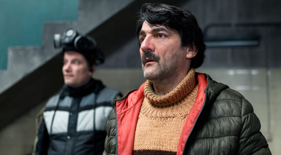 Auf dem Bild zur Serie Eternauta sind zwei Männer in einem Innenraum mit kühler Beton-Atmosphäre zu sehen. Rechts steht Favalli, gespielt von César Troncoso, mit ernstem Gesichtsausdruck, orangefarbenem Rollkragenpullover und olivgrüner Jacke. Im Hintergrund links ist Lucas, gespielt von Marcelo Subiotto, mit Winterjacke und aufgesetzter Gasmaske zu erkennen. Die Stimmung wirkt angespannt und nachdenklich.