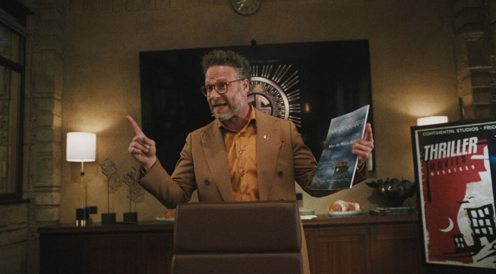 Auf dem Bild in dem Artikel zu The Studio Ende erklärt ist ein Charakter mittleren Alters in einem braunen Anzug und orangefarbenem Hemd zu sehen, wie er energisch spricht und dabei mit dem Finger gestikuliert. In der anderen Hand hält er ein Filmplakat. Im Hintergrund erkennt man ein modernes Büro.