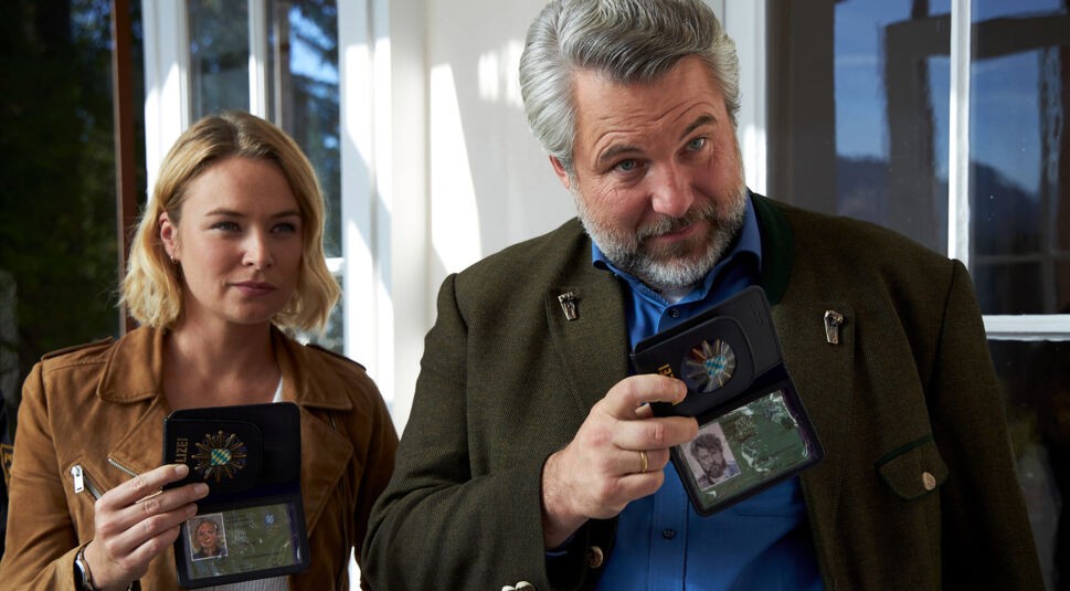 Im Bild zur ZDF Serien Empfehlungen sind Kommissarin Eva Winter, gespielt von Vanessa Eckart, und Kommissar Anton Stadler, gespielt von Dieter Fischer, bei einer Ermittlungsszene aus „Die Rosenheim-Cops“ zu sehen. Beide halten ihre Polizeiausweise in die Kamera, stehen in einem hellen Raum mit großen Fenstern und schauen konzentriert in Richtung einer Person außerhalb des Bildes.