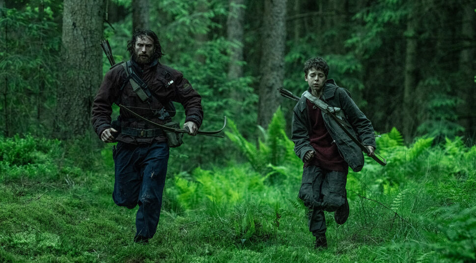 Auf dem Bild zur 28 Years Later-Kritik sind Jamie, gespielt von Aaron Taylor-Johnson, und Spike, dargestellt von Alfie Williams, in einer spannungsgeladenen Szene im dichten Wald zu sehen. Beide tragen dunkle Kleidung und Bögen, während sie durch das grüne Unterholz rennen. Die Atmosphäre wirkt düster und bedrohlich, passend zum post-apokalyptischen Setting des Films.