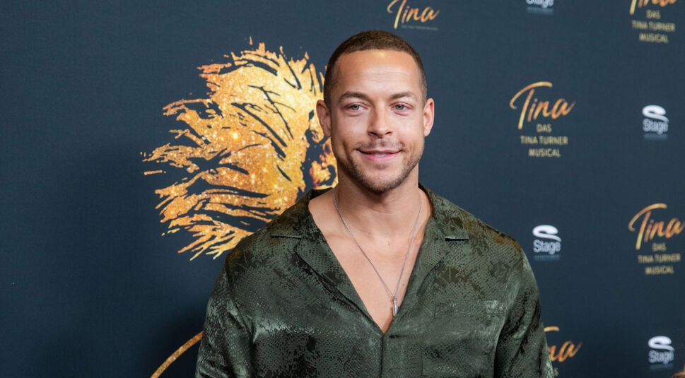 Auf dem Bild zur Übersicht Alle Bachelor ist Andrej Mangold vor einer Fotowand des Tina Turner Musicals zu sehen. Er trägt ein dunkelgrünes Hemd mit Schlangenledermuster und eine silberne Kette. Sein sympathisches Lächeln und sein sportlicher Look stehen sinnbildlich für seine Zeit als charismatischer Bachelor, der bei den Fans besonders beliebt war.