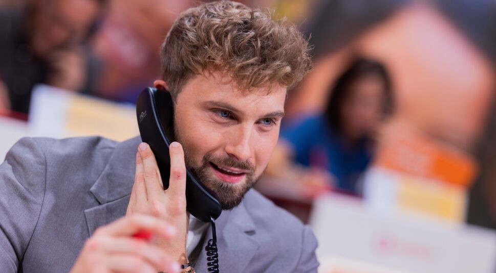 Auf dem Bild zur Übersicht Alle Bachelor ist Dominik Stuckmann im Einsatz bei einem Spenden- oder Charity-Event zu sehen. Er trägt ein graues Sakko und hält einen Telefonhörer am Ohr, während er konzentriert und engagiert wirkt. Sein Look ist modern und gepflegt – ganz im Stil des Bachelors, der als bodenständig und empathisch wahrgenommen wurde.