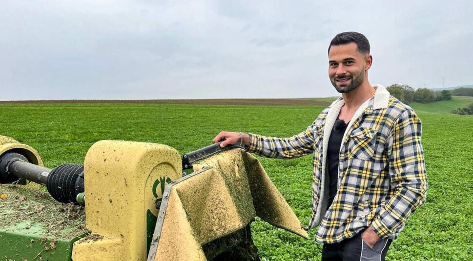 Bei Bauer sucht Frau 2025 steht Kandidat Basti in kariertem Hemd lässig neben einer landwirtschaftlichen Maschine auf einem weitläufigen grünen Feld und lächelt in die Kamera.
