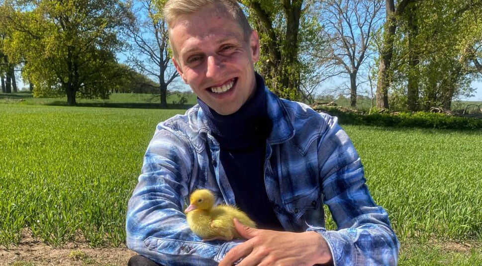 Bei Bauer sucht Frau 2025 sitzt Kandidat Christopher mit einem kleinen gelben Entenküken auf dem Arm lächelnd auf einem grünen Feld. Die Szene wirkt warmherzig und naturverbunden.