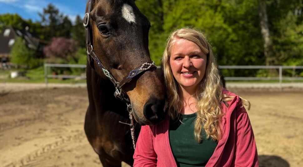 Bei Bauer sucht Frau 2025 steht Kandidatin Insa mit blondem Haar und roter Jacke neben einem braunen Pferd auf dem Reitplatz. Sie lächelt offen in die Kamera bei strahlendem Sonnenschein.